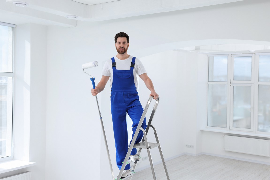 Handyman with roller on step ladder in room. Ceiling painting